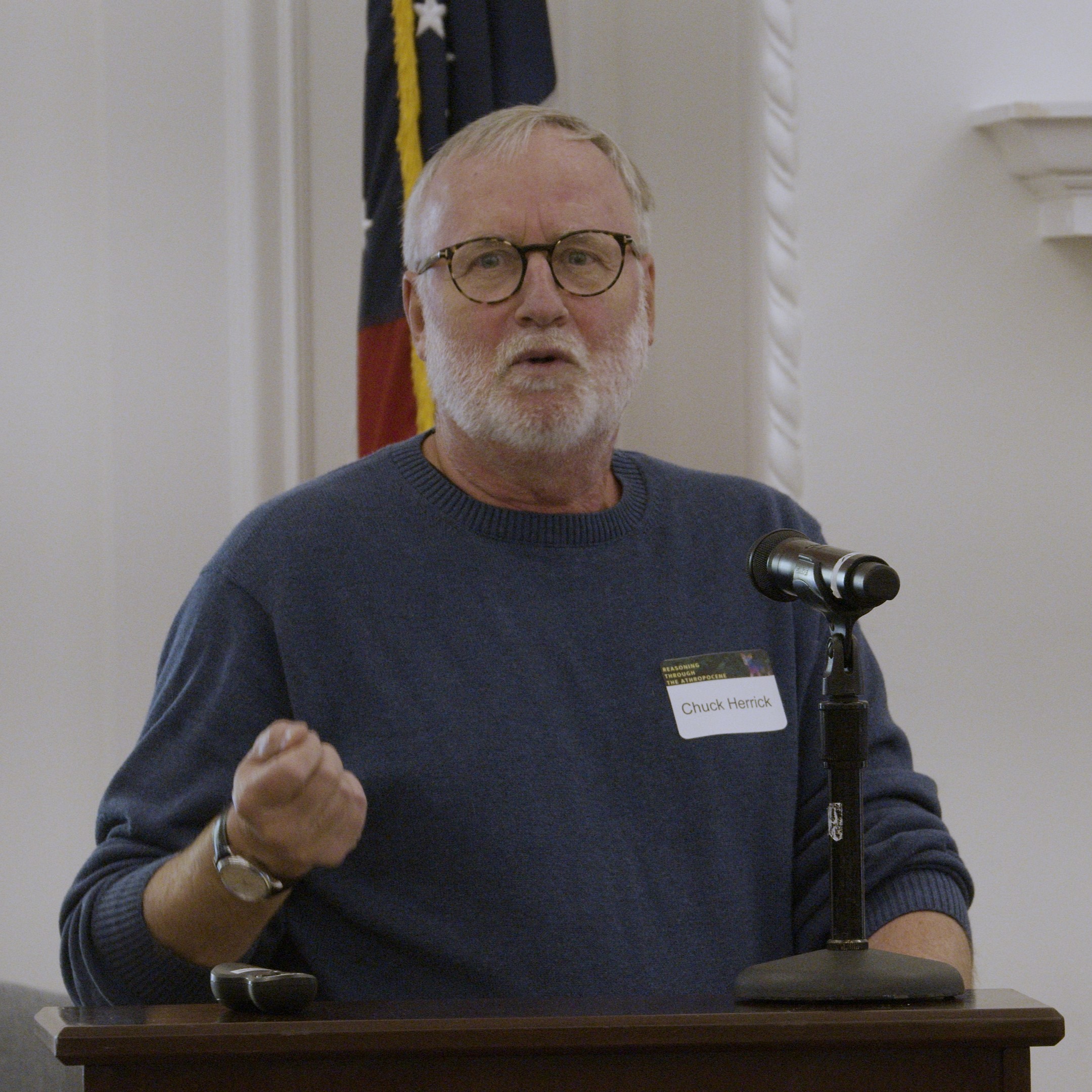 Photo of man in sweater presenting at a podium with an american flag behind him